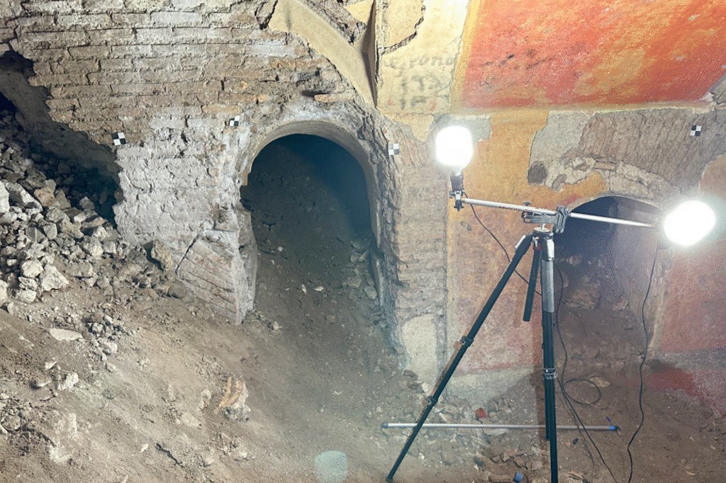 Interior de uma escava&ccedil;&atilde;o arqueol&oacute;gica com duas aberturas em arco na parede de tijolos e terra, iluminadas por um trip&eacute; com duas luzes fortes. O ch&atilde;o &eacute; de terra e pedras soltas, e a parede superior tem vest&iacute;gios de pintura avermelhada e amarelada