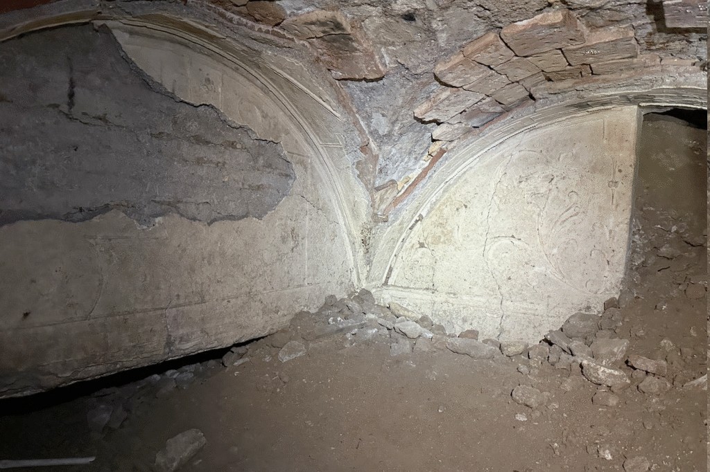 Interior de uma estrutura antiga com teto abobadado de tijolos e paredes de argamassa desbotada. H&aacute; duas ab&oacute;badas com detalhes em relevo e parte do reboco descascado, revelando a estrutura interna. O ch&atilde;o &eacute; de terra com entulho de pedras e argamassa, sugerindo um local em ru&iacute;nas ou em escava&ccedil;&atilde;o