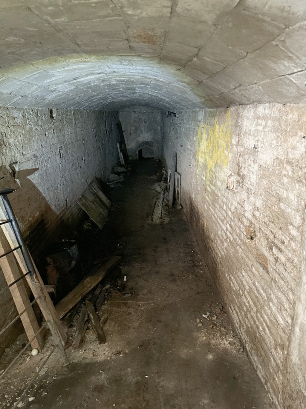 Corredor subterr&acirc;neo escuro e &uacute;mido com teto abobadado de tijolos. As paredes de tijolos brancos est&atilde;o descascadas e manchadas, com lixo e t&aacute;buas de madeira espalhados pelo ch&atilde;o de terra batida. Uma mancha amarela vibrante se destaca na parede direita, enquanto o fundo do corredor &eacute; quase totalmente escuro, com uma pequena abertura arqueada vis&iacute;vel