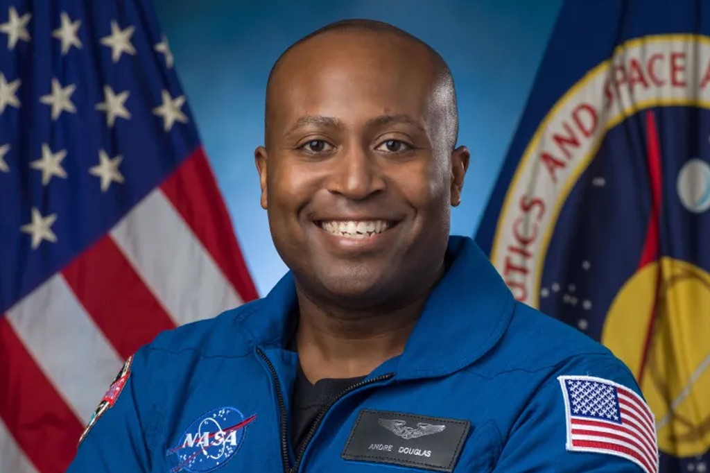 Homem negro careca sorrindo, vestindo uniforme azul da NASA, com bandeira dos EUA e logotipo da ag&ecirc;ncia espacial ao fundo