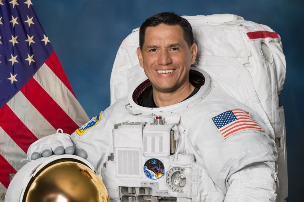 Astronauta sorridente, de pele morena e cabelo escuro, vestindo um traje espacial branco com a bandeira dos EUA no bra&ccedil;o, posa com um capacete dourado na m&atilde;o, em frente a uma bandeira americana e fundo azul