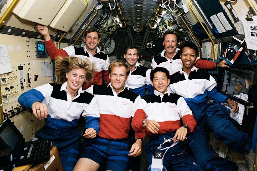 Sete astronautas sorridentes, quatro homens e tr&ecirc;s mulheres, flutuam dentro de uma espa&ccedil;onave, vestindo uniformes de polo brancos com faixas azuis e vermelhas. Eles posam para a c&acirc;mera, cercados por pain&eacute;is de controle e equipamentos da nave