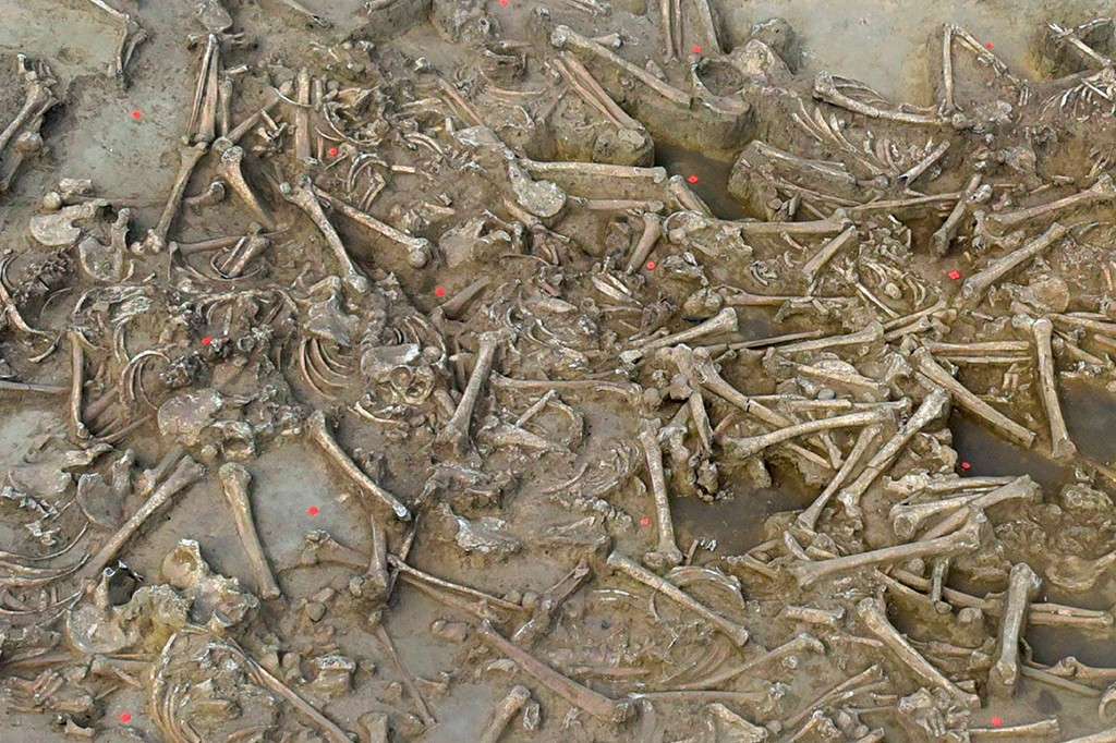 Vista aérea de uma vala comum com dezenas de esqueletos humanos desarticulados e ossos espalhados em solo úmido e marrom, com pontos vermelhos marcando alguns achados arqueológicos