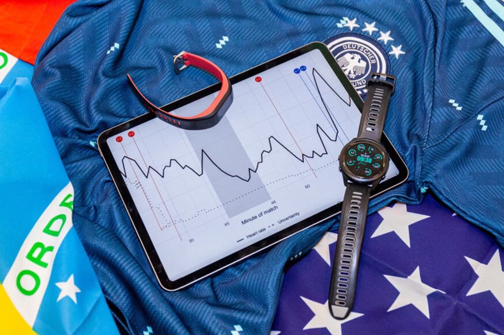 Tablet com gráfico de batimentos cardíacos durante uma partida, um relógio inteligente e uma pulseira fitness sobre uma camisa de futebol da Alemanha e bandeiras do Brasil e EUA