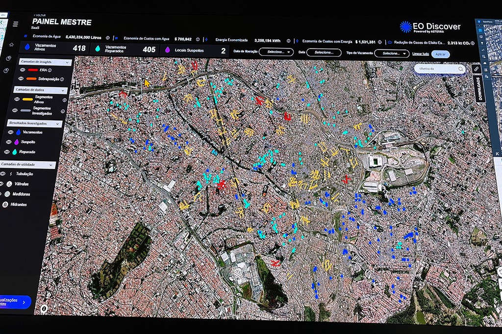 Tela de computador exibindo um mapa de sat&eacute;lite de uma cidade com &aacute;reas coloridas em vermelho, azul, amarelo e ciano, indicando vazamentos de &aacute;gua ativos e reparados, locais suspeitos e outras informa&ccedil;&otilde;es. No topo, um painel com dados como Vazamentos Ativos 418 e Vazamentos Reparados 405. &Agrave; esquerda, um menu com Camadas de insights, Camadas de dados, Resultados investigados e Camadas de utilidade. No canto superior direito, o logo EO Discover Powered by ASTERRA