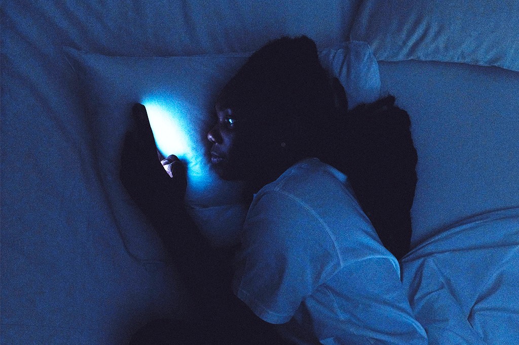 Pessoa deitada na cama no escuro, com o rosto iluminado pela luz azul de um celular que segura com a mão esquerda