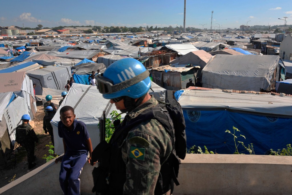 Um soldado da ONU com capacete azul e óculos de proteção, vestindo uniforme camuflado com a bandeira do Brasil no braço, observa um acampamento de refugiados. Tendas de lona cinza e azul se estendem sob um céu claro, com um menino de camisa azul olhando para a câmera em primeiro plano. Ao fundo, mais tendas e algumas construções precárias.