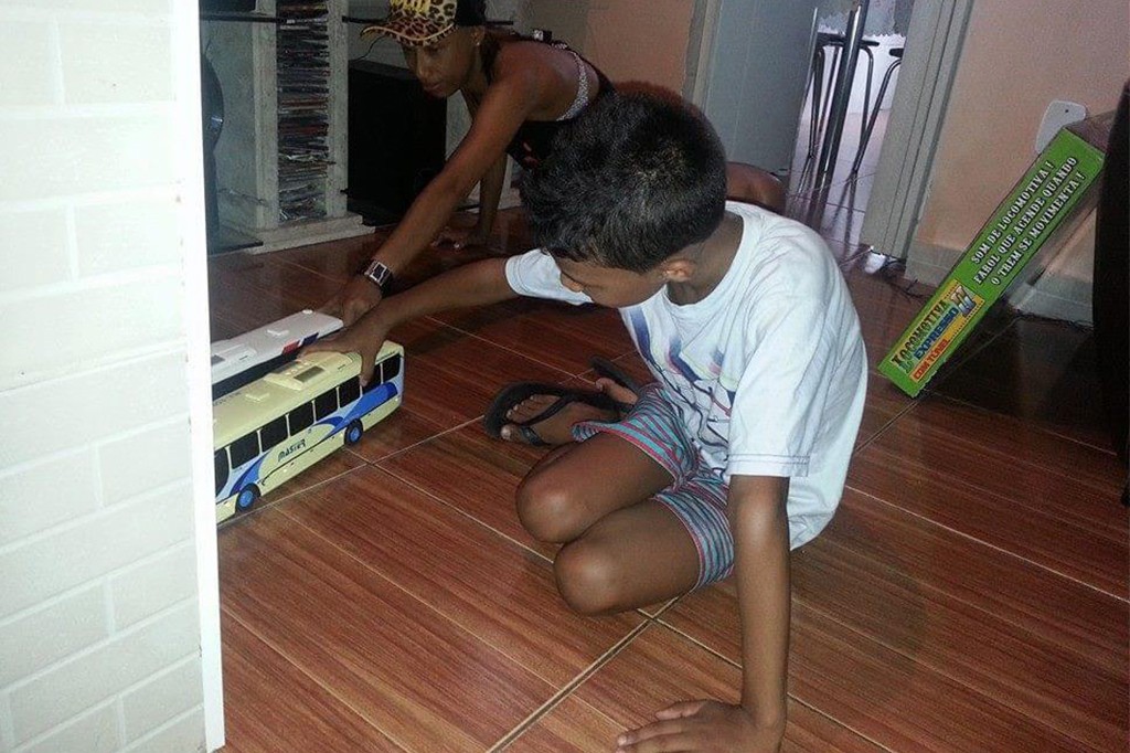 Duas crian&ccedil;as brincam no ch&atilde;o de madeira, um menino de camiseta branca e shorts listrados empurra um &ocirc;nibus de brinquedo bege e azul, enquanto uma menina de bon&eacute; de oncinha e regata preta o ajuda. Ao fundo, uma caixa verde de Locomotiva Expresso com texto SOM DE LOCOMOTIVA! FAROL QUE ACENDE QUANDO O TREM SE MOVIMENTA!