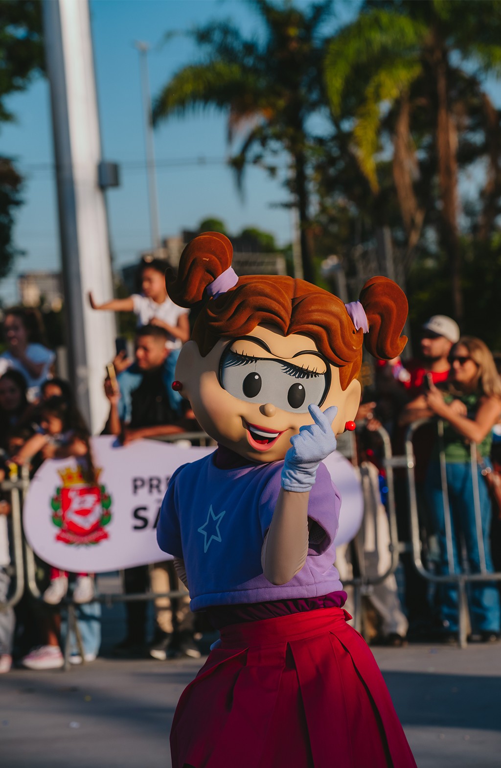 Mônica, personagem da Turma da Mônica, fantasiada com cabelo castanho e laços lilás, blusa lilás com estrela branca e saia vermelha, sorri e faz um coração com os dedos. Ela está em um evento ao ar livre, com pessoas e palmeiras ao fundo