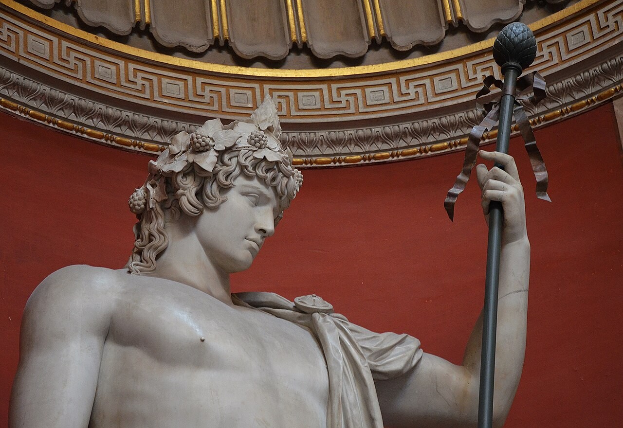 Est&aacute;tua de m&aacute;rmore de um homem jovem, com coroa de folhas e uvas, segurando um bast&atilde;o com uma pinha na ponta e fitas, contra uma parede vermelha e teto dourado com padr&otilde;es geom&eacute;tricos
