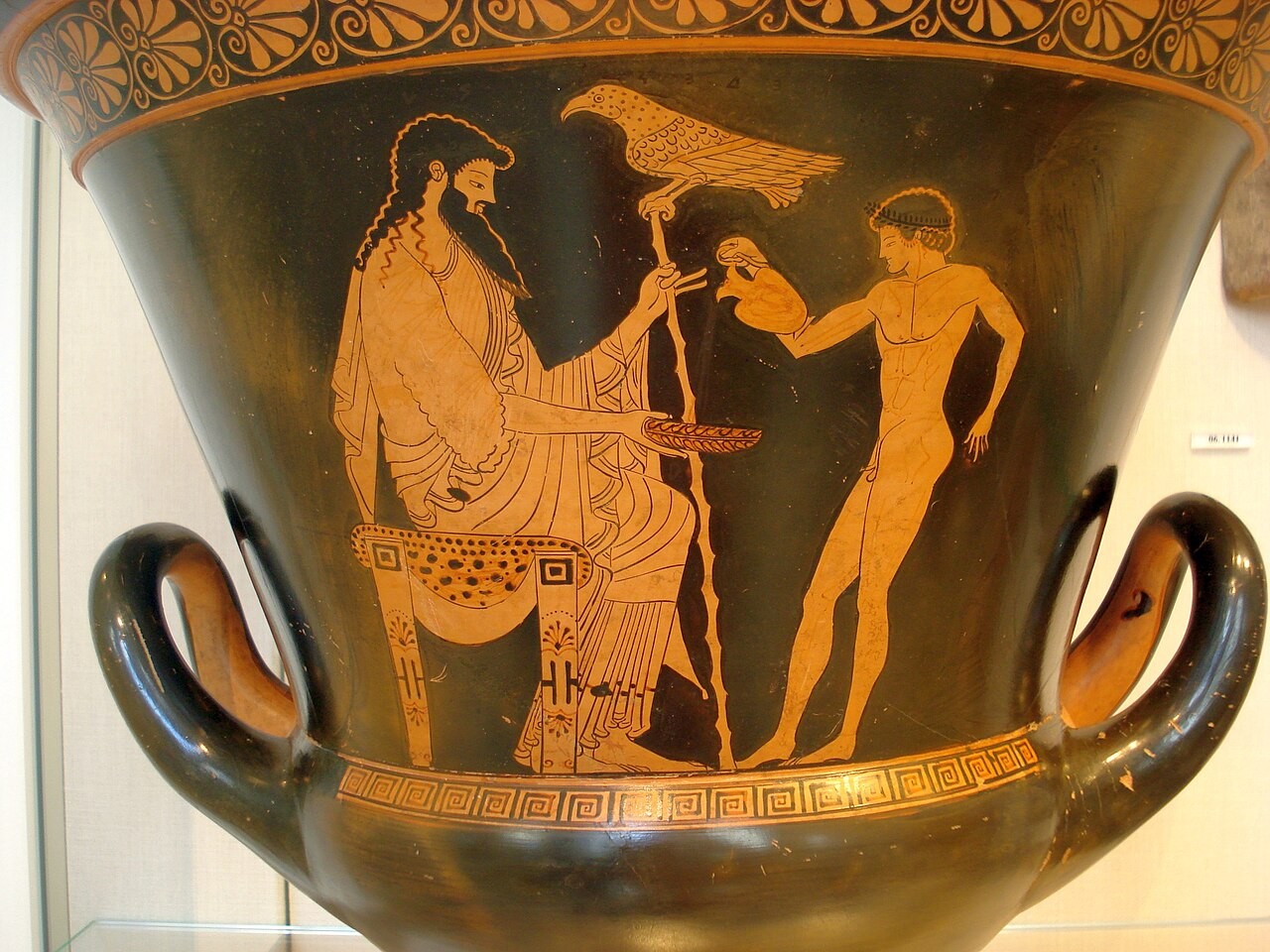 Pintura em vaso grego antigo, com fundo preto e figuras em terracota. Um homem barbudo e vestido senta-se &agrave; esquerda, segurando um cajado. Um p&aacute;ssaro pousa no cajado. &Agrave; direita, um jovem nu oferece um objeto ao homem sentado. Al&ccedil;as laterais e padr&otilde;es geom&eacute;tricos adornam o vaso