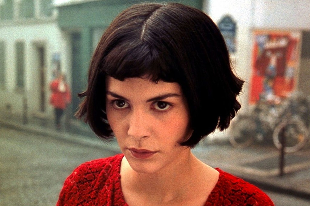 Mulher de cabelo curto e escuro, com franja reta, veste um su&eacute;ter vermelho texturizado e olha diretamente para frente com express&atilde;o s&eacute;ria. Ao fundo, uma rua borrada com edif&iacute;cios claros e uma pessoa de casaco vermelho distante.