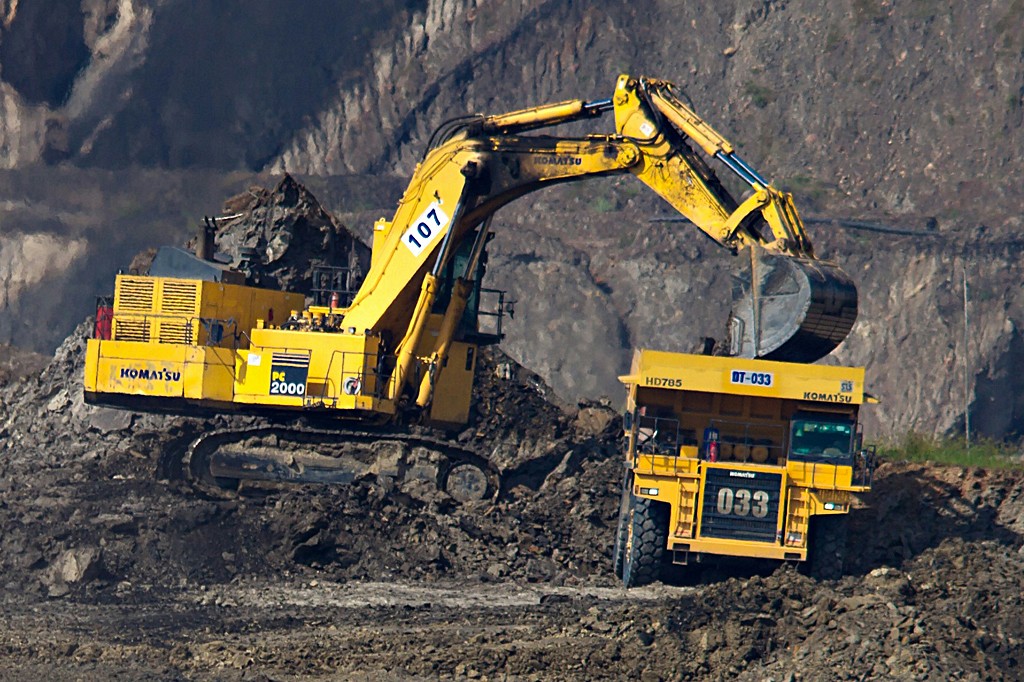 Escavadeira amarela Komatsu PC 2000 com número 107 no braço, carregando terra em um caminhão basculante Komatsu HD785 com número 033, em uma mina a céu aberto.