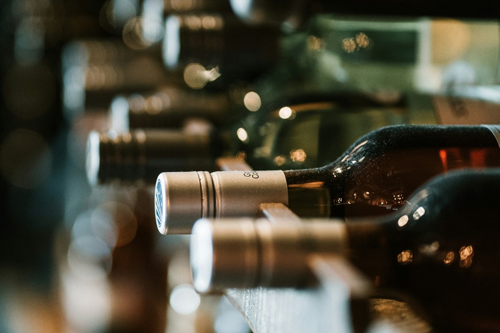 Garrafas de vinho escuras, com rótulos e tampas prateadas, alinhadas horizontalmente em uma adega escura, com pontos de luz desfocados ao fundo.