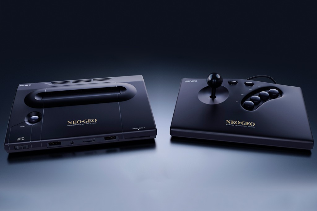 Console e controle Neo-Geo pretos, com detalhes dourados, sobre uma superfície escura e reflexiva. O console está à esquerda, com botões Reset e Power On-Off. O controle, à direita, possui um joystick e quatro botões redondos pretos.