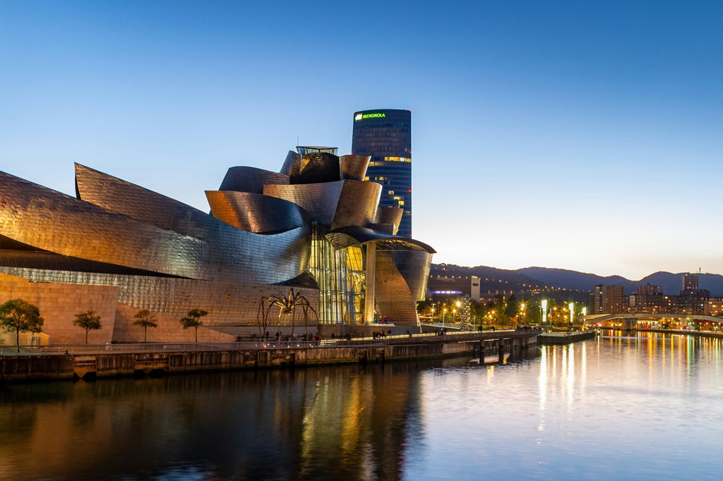 Imagem do Museu Guggenheim Bilbao, com sua arquitetura metálica curvilínea, reflete a luz do entardecer sobre o rio. Ao fundo, um arranha-céu e a cidade iluminada se estendem até as montanhas. Uma escultura de aranha gigante está em frente ao museu.