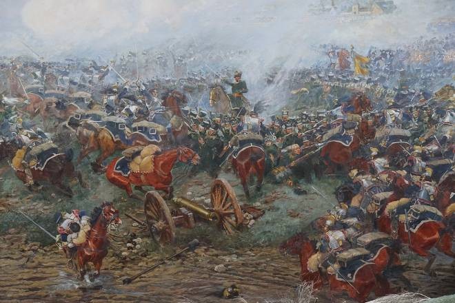 Pintura de uma batalha histórica com muitos soldados a cavalo e a pé, em meio a fumaça e poeira. Canhões e armas de fogo são visíveis no campo de batalha, com um castelo ao fundo.