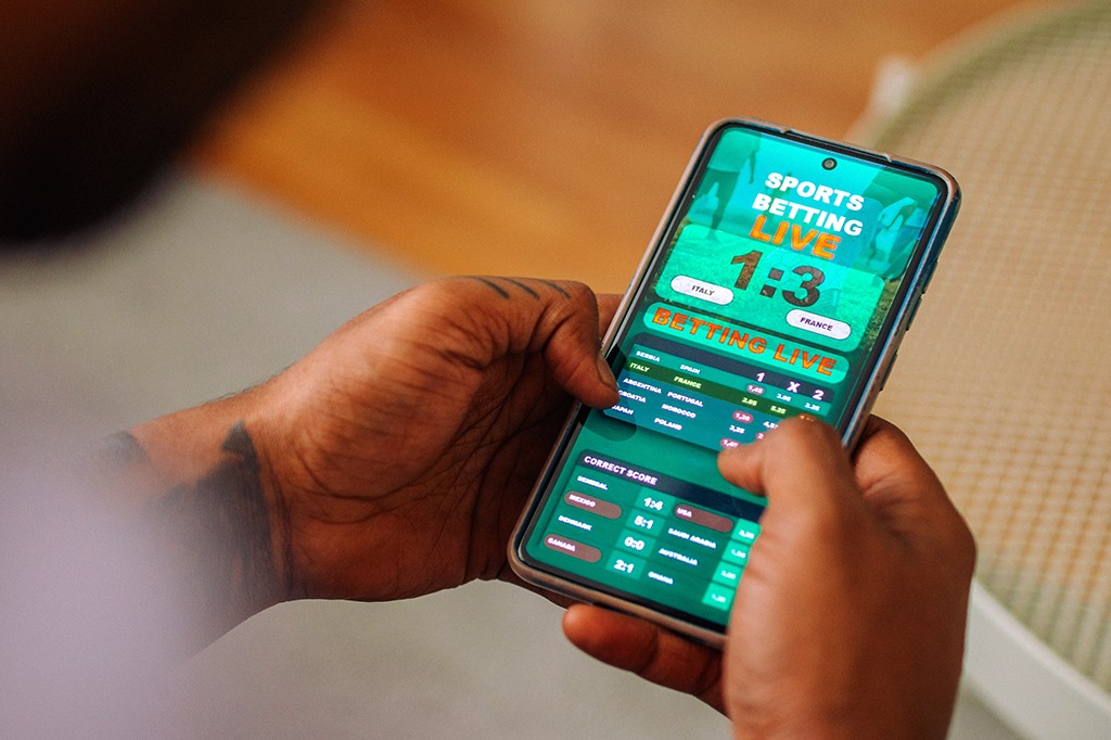 Mãos de uma pessoa com tatuagens segurando um smartphone que exibe um aplicativo de apostas esportivas ao vivo, com placar de 1:3 entre Itália e França, e outras opções de apostas