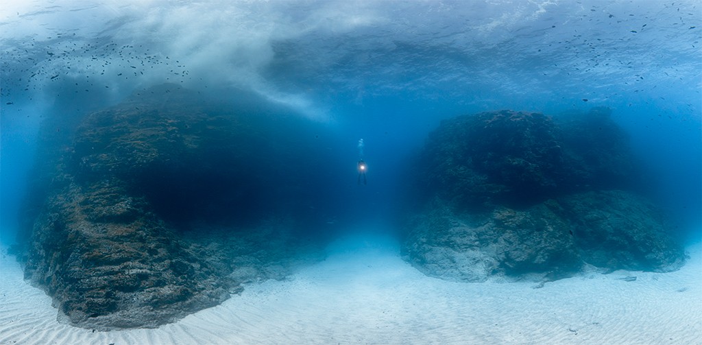 Mergulhador solit&aacute;rio com lanterna ilumina o fundo do mar azul profundo, entre duas grandes forma&ccedil;&otilde;es rochosas escuras e um leito de areia clara. Pequenos peixes nadam perto da superf&iacute;cie.