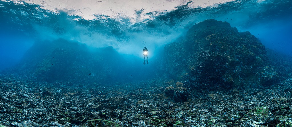 Mergulhador solit&aacute;rio com uma lanterna subaqu&aacute;tica brilhante, nadando em &aacute;guas azuis profundas sobre um leito marinho rochoso e irregular. A superf&iacute;cie da &aacute;gua, acima, reflete a luz do sol em ondas. &Agrave; direita, uma grande forma&ccedil;&atilde;o rochosa escura se eleva do fundo.