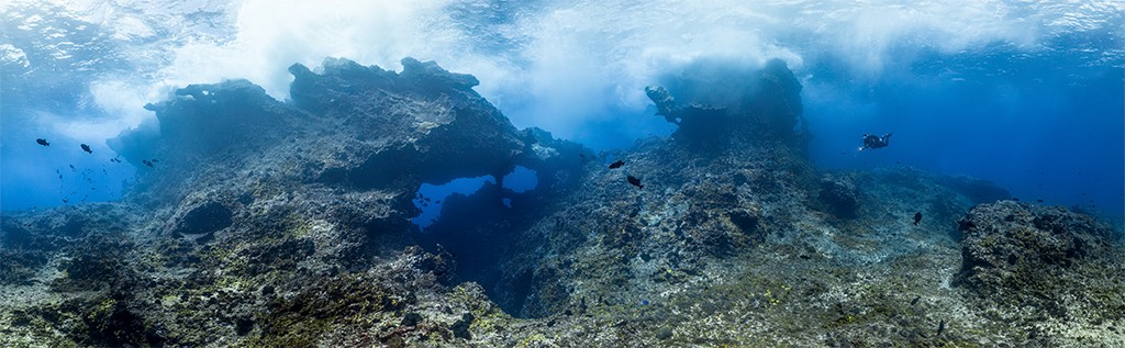 Vista subaqu&aacute;tica de um recife de coral rochoso com forma&ccedil;&otilde;es escuras e irregulares, algumas com aberturas azuis. Pequenos peixes escuros nadam perto das rochas. No canto superior direito, um mergulhador solit&aacute;rio explora a &aacute;gua azul-clara, com a superf&iacute;cie ondulada vis&iacute;vel acima