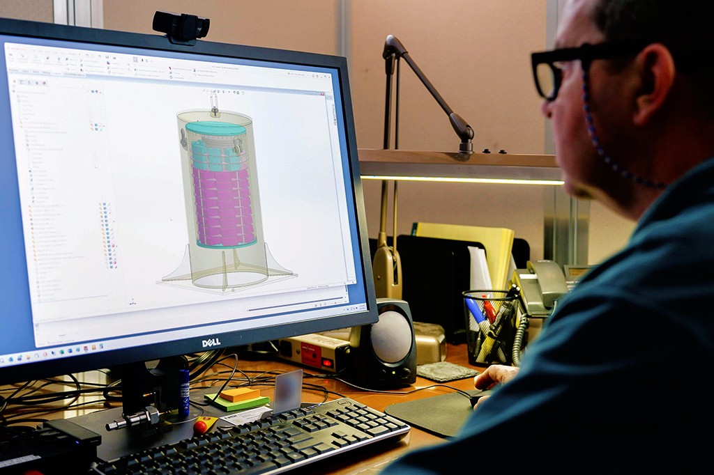 Homem de &oacute;culos e camisa azul, de costas, usando um computador Dell com um software de design 3D exibindo um cilindro transparente com partes internas azuis e roxas. A mesa tem teclado, mouse, alto-falante, telefone e porta-canetas.