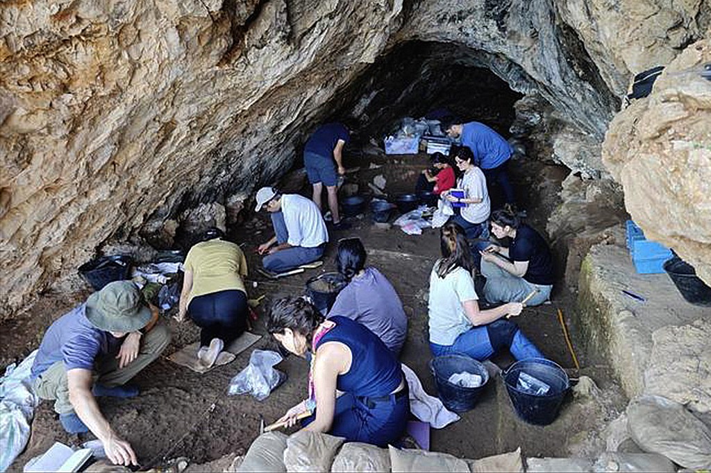 Arque&oacute;logos, homens e mulheres, escavam cuidadosamente o ch&atilde;o de uma caverna rochosa, usando ferramentas pequenas e baldes, em um s&iacute;tio arqueol&oacute;gico