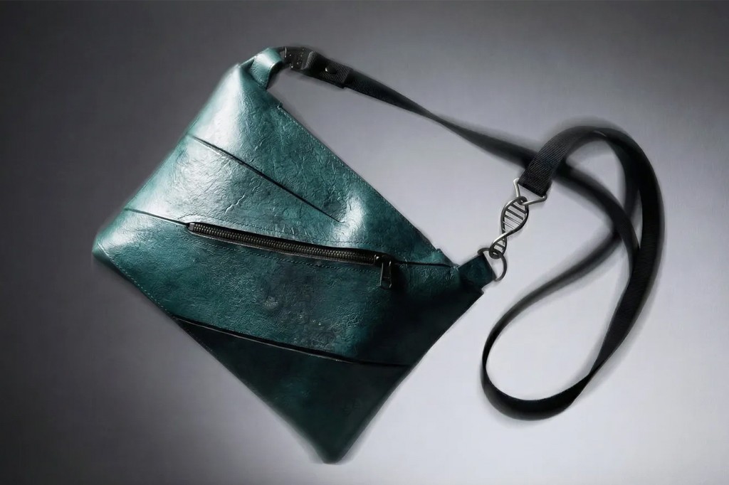 Bolsa transversal verde-escura com textura enrugada, z&iacute;per frontal preto e al&ccedil;a preta com um chaveiro em formato de dupla h&eacute;lice de DNA