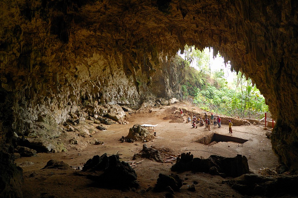 Interior de uma caverna ampla e escura, com teto e paredes rochosas irregulares, cheias de estalactites. No fundo, um grupo de arque&oacute;logos trabalha em escava&ccedil;&otilde;es, com a luz do dia e vegeta&ccedil;&atilde;o exuberante vis&iacute;veis na entrada da caverna