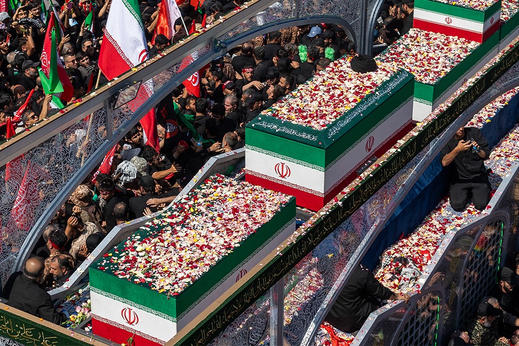 Multidão de pessoas vestindo preto e verde, algumas segurando bandeiras do Irã, acompanham caixões cobertos com a bandeira iraniana e pétalas de flores vermelhas e brancas, em um funeral ou procissão. Os caixões estão em uma estrutura elevada, e a multidão se aglomera ao redor e abaixo, com alguns indivíduos ajoelhados ou tocando os caixões