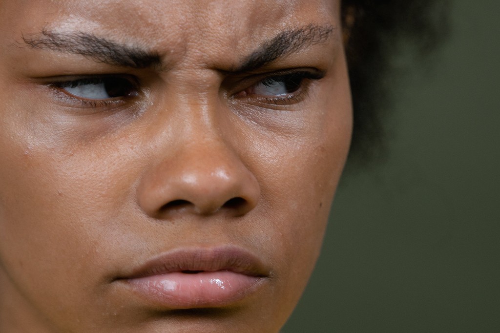 Rosto de uma mulher negra com expressão de desconfiança, sobrancelhas franzidas e lábios cerrados, olhando para a direita, em um fundo verde escuro
