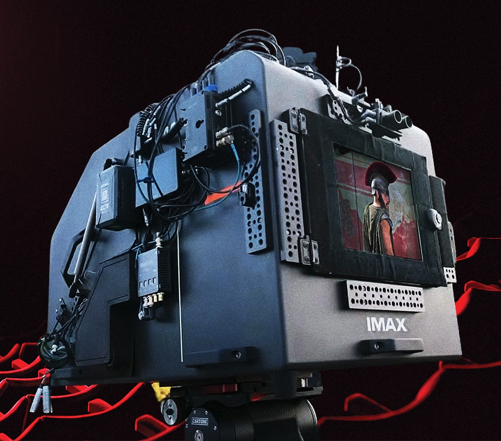 C&acirc;mera de cinema IMAX cinza escura com cabos e equipamentos conectados, exibindo um soldado romano de perfil em seu monitor. O fundo &eacute; de cadeiras vermelhas de cinema no fundo preto.