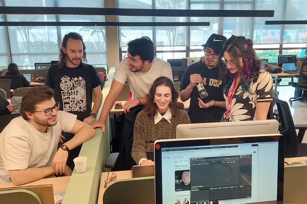 Seis jovens adultos, sorriem enquanto trabalham juntos em um escrit&oacute;rio moderno.