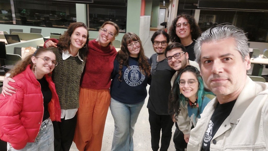 Grupo de nove pessoas sorrindo para a c&acirc;mera em um ambiente interno com mesas e cadeiras ao fundo. Todos parecem felizes e descontra&iacute;dos.