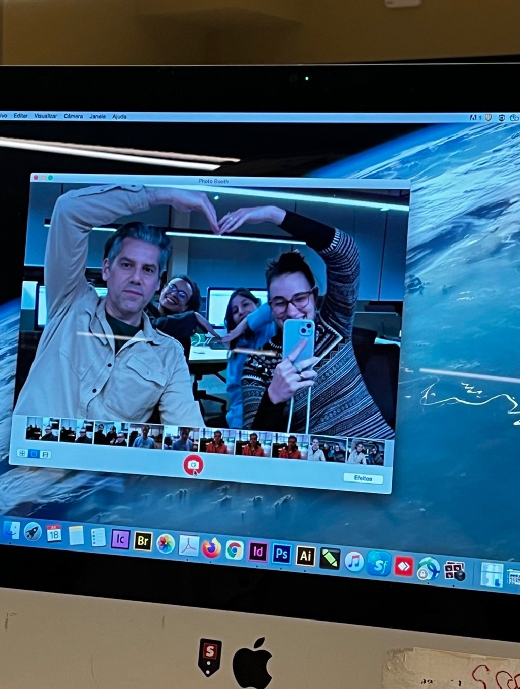 Quatro pessoas sorrindo, duas &agrave; frente fazendo um cora&ccedil;&atilde;o com as m&atilde;os, e duas ao fundo, completando o formato, s&atilde;o refletidas na tela de um computador com fundo de imagem da Terra