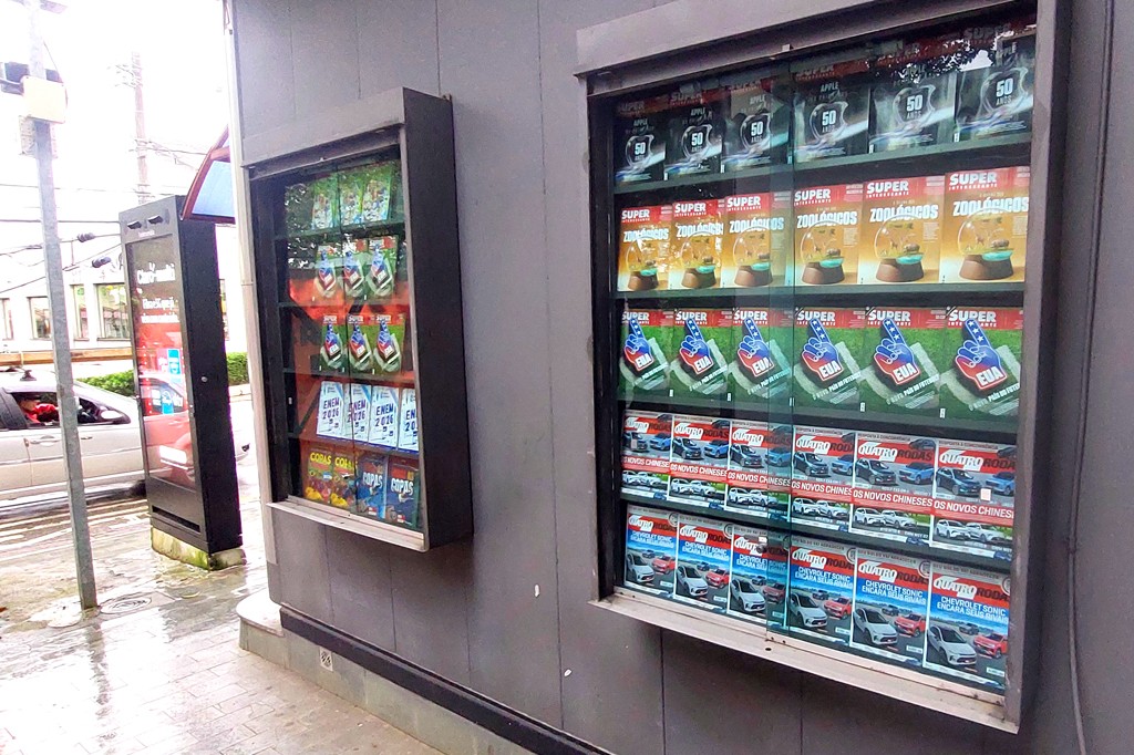Duas vitrines, de uma banca de jornal, de vidro exibem revistas. A vitrine maior &agrave; direita mostra tr&ecirc;s edi&ccedil;&otilde;es da revista Super Interessante e um da Quatro Rodas. A banca menor &agrave; esquerda v&ecirc;-se um Guia ENEM 2026 e Copas. O ch&atilde;o de cimento est&aacute; molhado, e um carro prateado passa ao fundo.