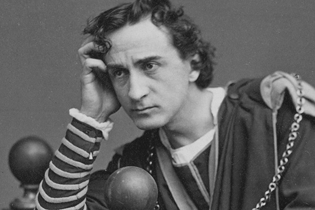Homem com cabelo encaracolado, vestindo t&uacute;nica escura e camisa listrada, apoia a cabe&ccedil;a na m&atilde;o direita, olhando pensativo para a frente. Duas esferas escuras e uma corrente met&aacute;lica aparecem no primeiro plano.