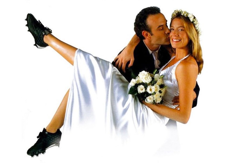 Um homem de terno preto beija a bochecha de uma mulher sorridente, vestida de noiva com um buqu&ecirc; de rosas brancas e uma coroa de flores. Ela est&aacute; sendo carregada, com uma perna estendida para cima, cal&ccedil;ando uma chuteira preta de futebol