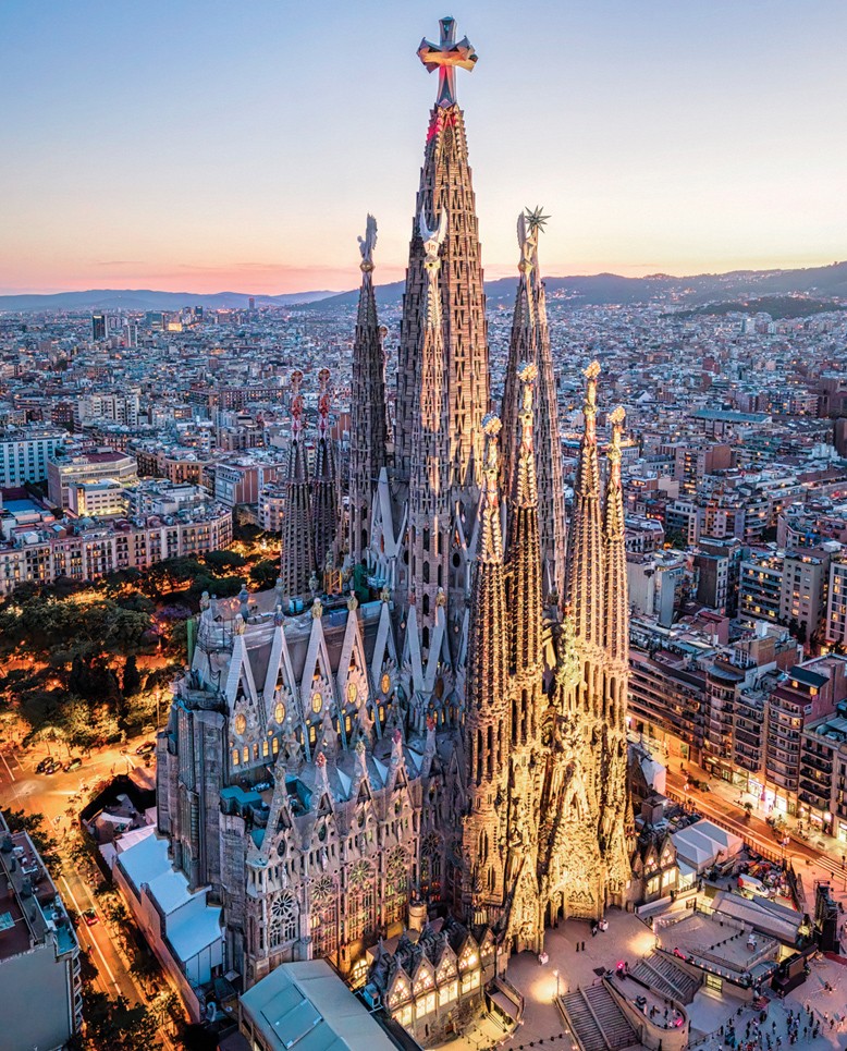 Vista a&eacute;rea da Bas&iacute;lica da Sagrada Fam&iacute;lia em Barcelona, Espanha, ao entardecer. A arquitetura g&oacute;tica da igreja &eacute; iluminada por luzes quentes, destacando suas torres pontiagudas e detalhes intrincados. Ao fundo, a cidade se estende com edif&iacute;cios e ruas iluminadas, sob um c&eacute;u em tons de azul e rosa.