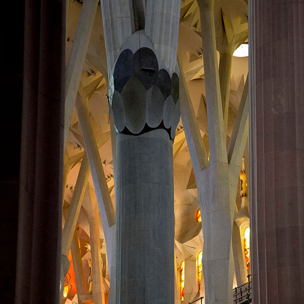 Interior da Sagrada Fam&iacute;lia com colunas ramificadas que se assemelham a &aacute;rvores, iluminadas por luzes quentes e vitrais coloridos. Uma coluna central cinza tem um capitel decorado com formas ovais escuras, e outras colunas claras se estendem para o teto, criando um ambiente grandioso e complexo