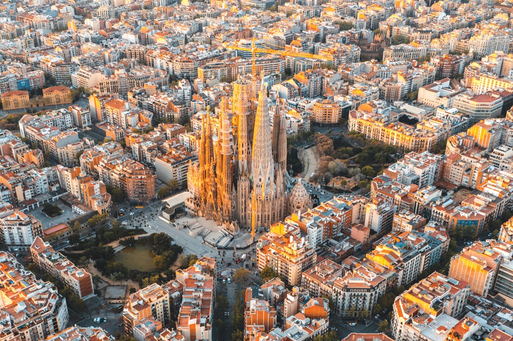 Vista a&eacute;rea da Sagrada Fam&iacute;lia em Barcelona, com suas torres g&oacute;ticas iluminadas pelo sol poente, cercada por quarteir&otilde;es de edif&iacute;cios residenciais e comerciais de cor clara, com telhados alaranjados e ruas movimentadas. H&aacute; um parque com lago &agrave; esquerda da bas&iacute;lica e guindastes de constru&ccedil;&atilde;o vis&iacute;veis ao fundo, indicando a expans&atilde;o urbana