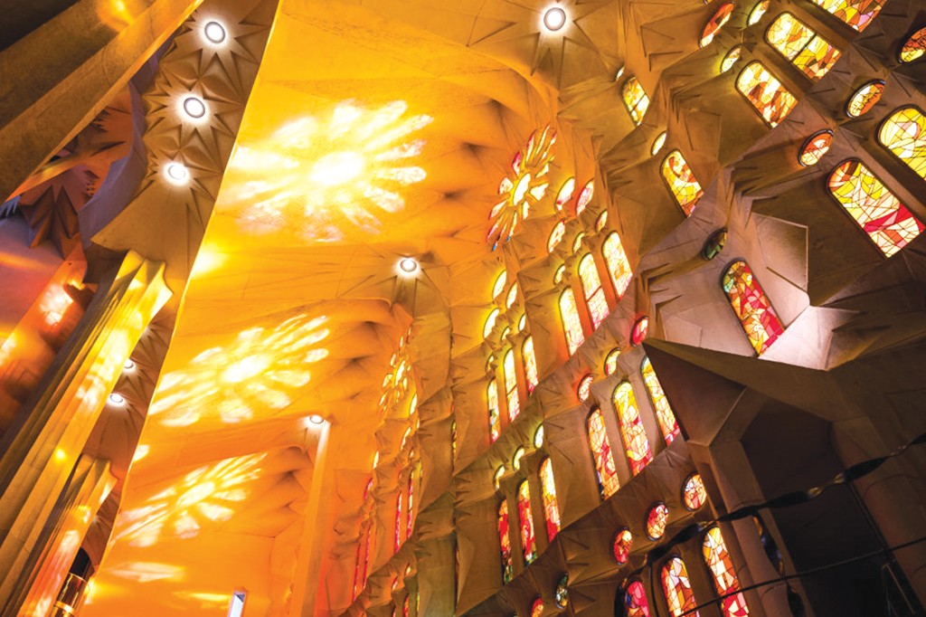 Interior de uma igreja com teto alto e abobadado, iluminado por luzes circulares e proje&ccedil;&otilde;es de luz em formato de estrela. &Agrave; direita, uma parede com m&uacute;ltiplos vitrais coloridos em tons de vermelho, laranja e amarelo, criando um ambiente quente e vibrante