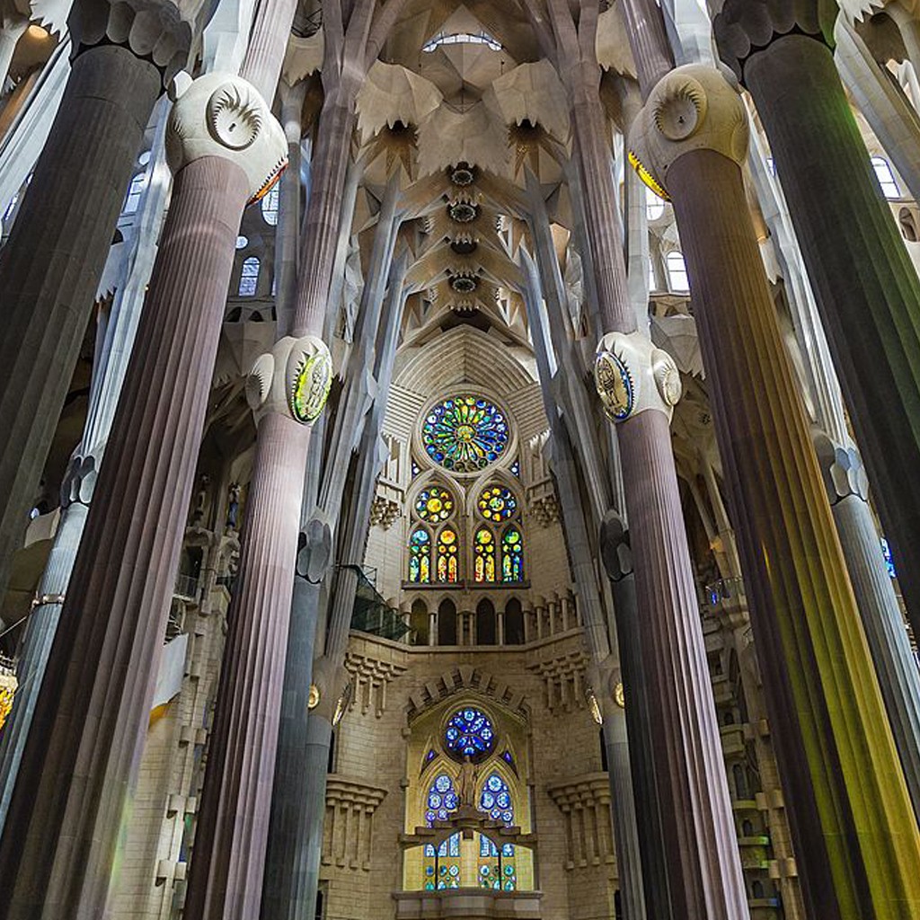 Interior da Bas&iacute;lica da Sagrada Fam&iacute;lia, com colunas altas e ramificadas que se assemelham a &aacute;rvores, sustentando um teto abobadado. H&aacute; vitrais coloridos em formato circular e retangular, iluminando o ambiente com tons vibrantes de azul, verde, amarelo e vermelho. A arquitetura g&oacute;tica modernista &eacute; grandiosa e detalhada
