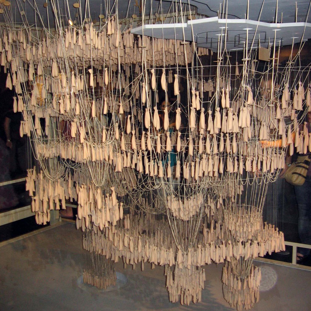 Instala&ccedil;&atilde;o art&iacute;stica com centenas de pequenos sacos de tecido bege pendurados por fios finos, formando uma estrutura densa e org&acirc;nica que se estende do teto ao ch&atilde;o, com pessoas observando ao fundo