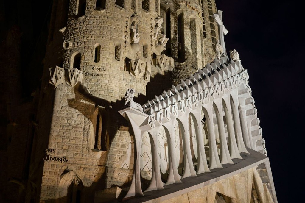 Fachada da Sagrada Fam&iacute;lia &agrave; noite, com ilumina&ccedil;&atilde;o destacando a torre de pedra e uma estrutura branca com arcos e pin&aacute;culos triangulares. Esculturas e inscri&ccedil;&otilde;es latinas como Corda Surcum e Sus Joseph s&atilde;o vis&iacute;veis, contra um c&eacute;u escuro