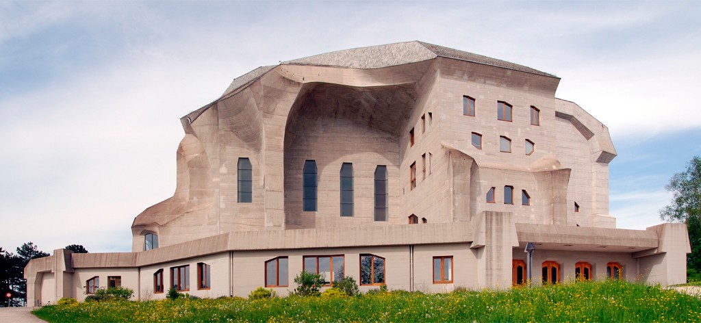 Edif&iacute;cio de concreto com arquitetura brutalista, formas org&acirc;nicas e telhado irregular, janelas retangulares e arqueadas, cercado por gramado verde com flores amarelas sob c&eacute;u azul claro