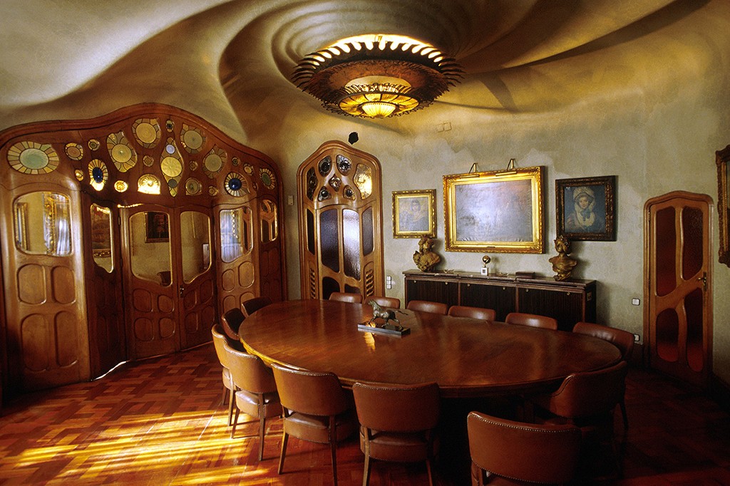 Sala de jantar com teto ondulado e lustre circular. Uma mesa oval de madeira escura com cadeiras de couro marrom ocupa o centro. Na parede, portas de madeira ornamentadas com vidros e espelhos, e quadros com molduras douradas. O ch&atilde;o de parquet recebe raios de sol