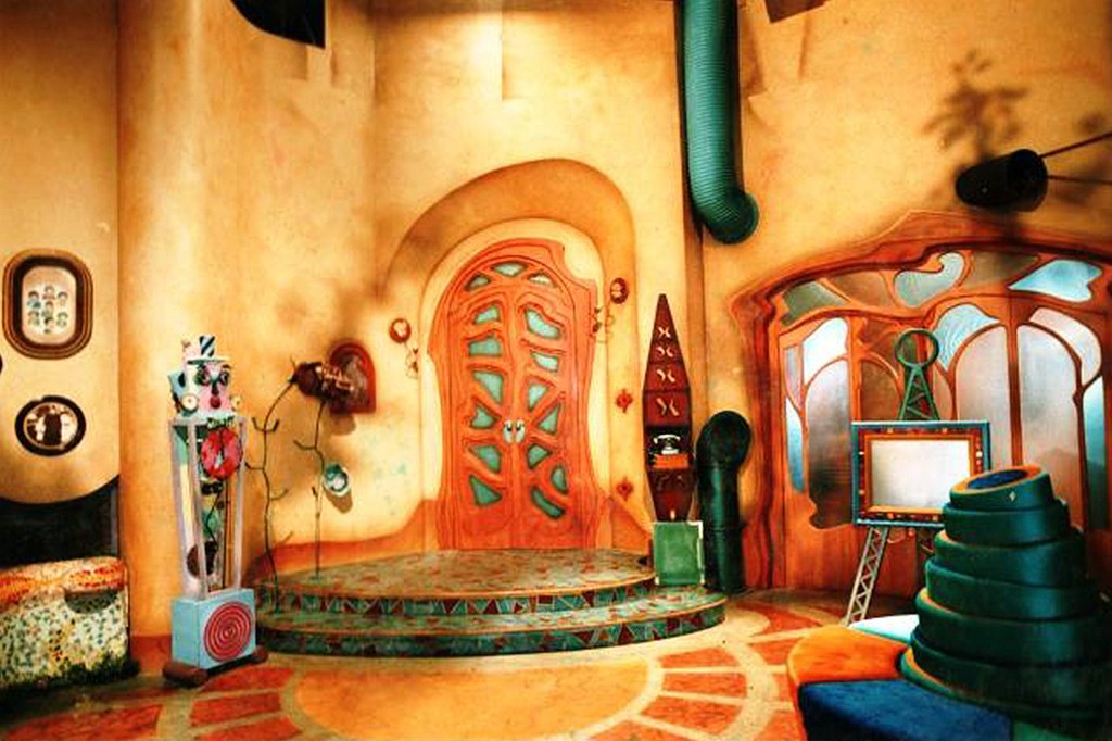 Cenário colorido e lúdico com paredes em tons de pêssego, porta de madeira entalhada em formato orgânico, degraus semicirculares com mosaicos azuis e verdes, e objetos decorativos excêntricos como um robô e uma televisão antiga