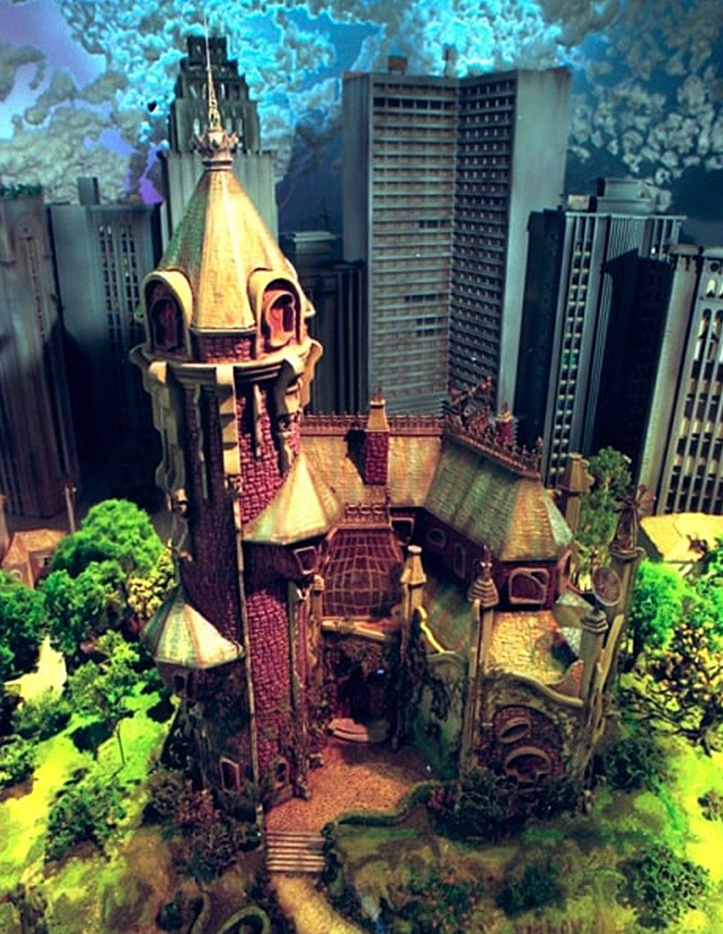 Maquete de um castelo g&oacute;tico com telhados verdes e paredes avermelhadas, cercado por &aacute;rvores e arbustos verdes, em contraste com arranha-c&eacute;us cinzentos ao fundo sob um c&eacute;u azul nublado