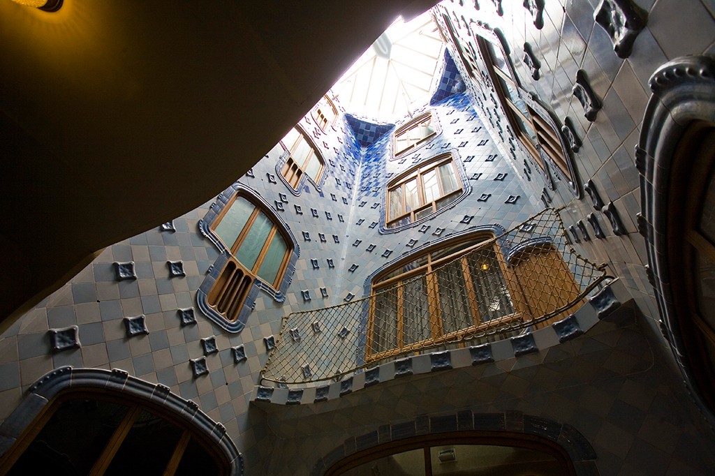 Vista de baixo para cima do p&aacute;tio interno da Casa Batll&oacute;, com paredes curvas revestidas de azulejos azuis e brancos, janelas de madeira de formatos org&acirc;nicos e um telhado de vidro que ilumina o ambiente